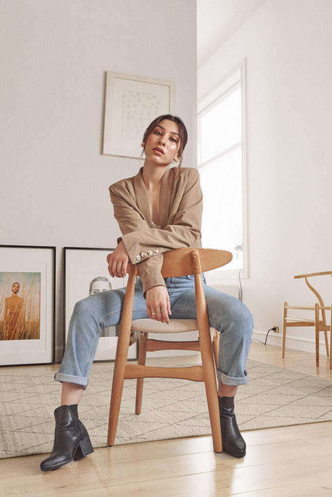 Nadya Inzunza is shown seated on a wooden chair in a minimalist room with framed artworks on the wall. She wears a beige blazer blue jeans and black ankle boots.